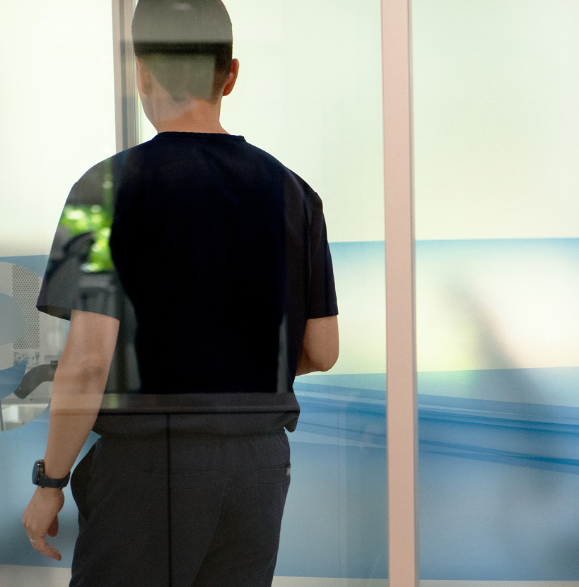 A person in a navy outfit is seen from behind through a glass window. The setting appears calm, with soft light and minimalistic decor in blue tones.